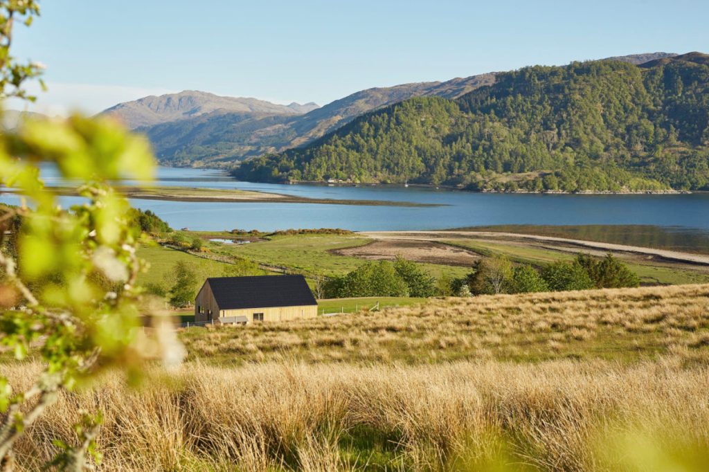 A lone Scandinavian-looking cabin from 57 Nord contemplates its life choices above a Scottish loch, surrounded by smugly photogenic hills pretending it’s always this sunny.