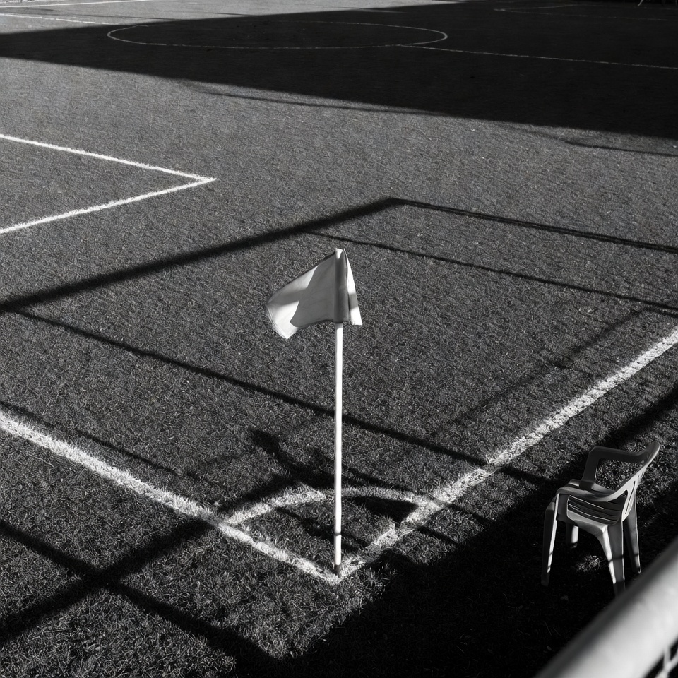 Black-and-white photograph of an empty, frost-dusted sports pitch at dawn, with long shadows, metal goalposts, and stacked plastic chairs, conveying the quiet absence of routine and community structure.