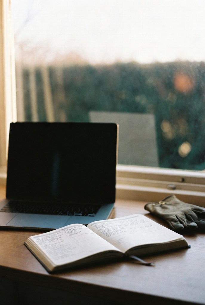 Minimal home office desk in soft winter light, with a laptop, an open weekly diary filled with handwritten notes, and worn leather gardening gloves beside a window overlooking a blurred garden hedge.